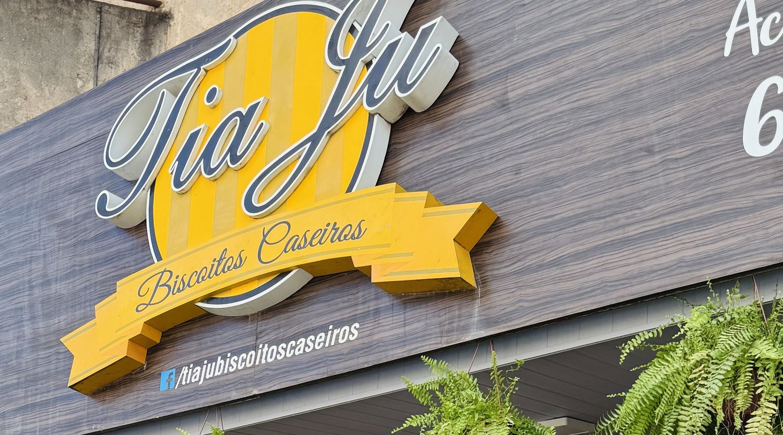 Fachada da padaria Tia Ju Biscoitos Caseiros, com letreiro amarelo e plantas na entrada