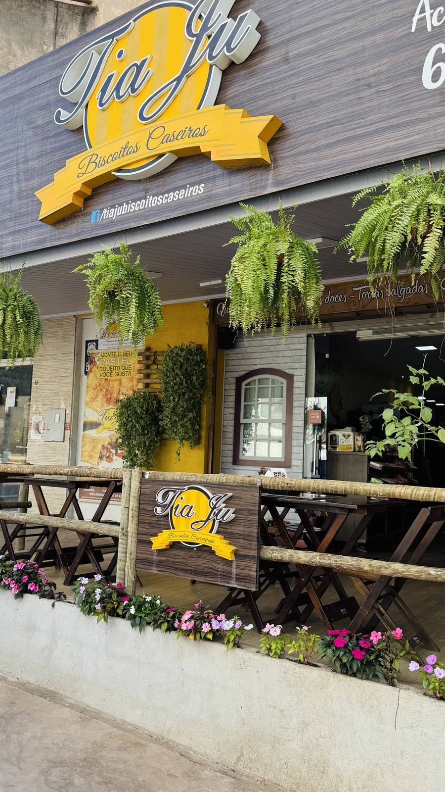 Fachada da padaria Tia Ju Biscoitos Caseiros, com letreiro amarelo e plantas na entrada